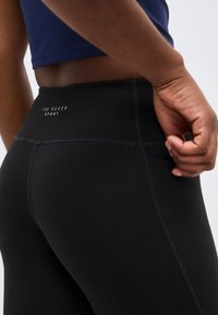 Κοντινό πλάνο ενός ατόμου που φοράει μαύρα ψηλόμεσα κολάν με το λογότυπο «Ted Baker Sport», χέρι ακουμπάει κοντά στην τσέπη του γοφού και ναυτικό μπλε cropped top.