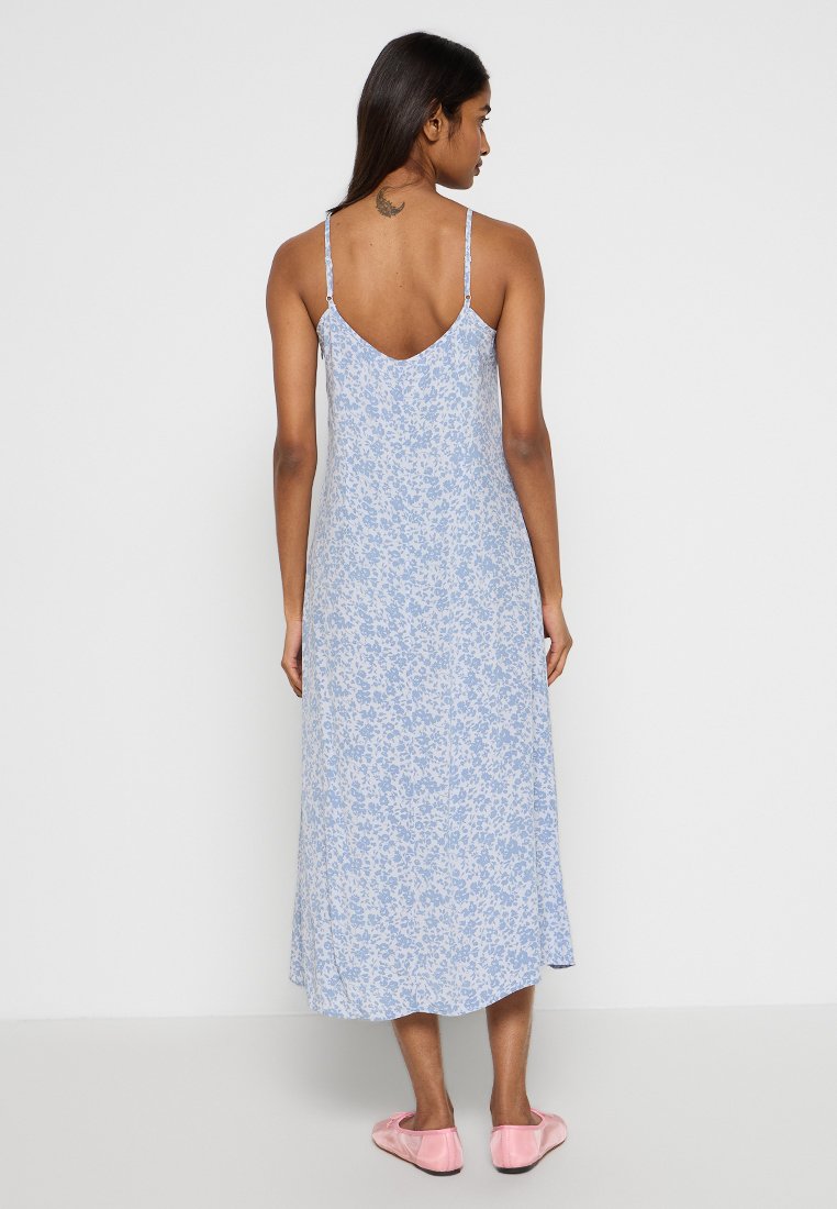 Frau steht mit dem Rücken zur Kamera, trägt ein hellblaues, blumiges Midikleid mit dünnen Trägern und rosa Satin-Ballettschuhe.