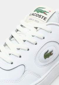 Witte leren sneaker met tonale stiksels, ronde neus en platte witte veters. Voorzien van een groen geborduurd krokodillenlogo aan de zijkant.
