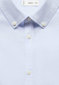 Lichtblauw shirt met een button-down kraag, met witte knopen en een gestructureerde stof. Label met "MNG" op de kraag.
