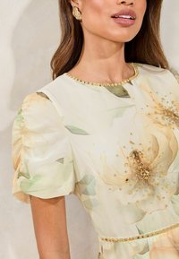 Abito floreale con colori pastello giallo e verde, maniche a sbuffo, dettagli in perline dorate sul colletto e decorazioni floreali sul busto.