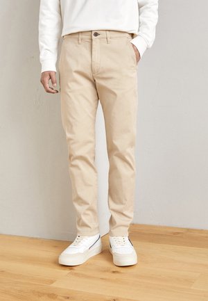 Man wearing beige slim-fit pants, white sneakers, and a white long-sleeve shirt standing on wood flooring against a plain wall.