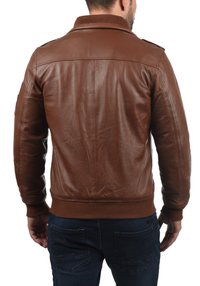 Bărbat îmbrăcat într-o geacă din piele maro și blugi bleu închis, întors cu spatele pe un fundal alb, arătând detaliile din spate ale gecii.