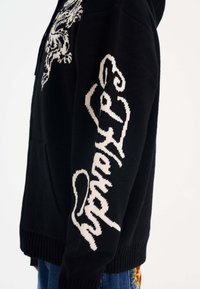 Felpa nera a maglia con cappuccio caratterizzata da grafiche bianche, tra cui una tigre e testo lungo la manica. Orlo e polsini a costine, con tasca frontale.