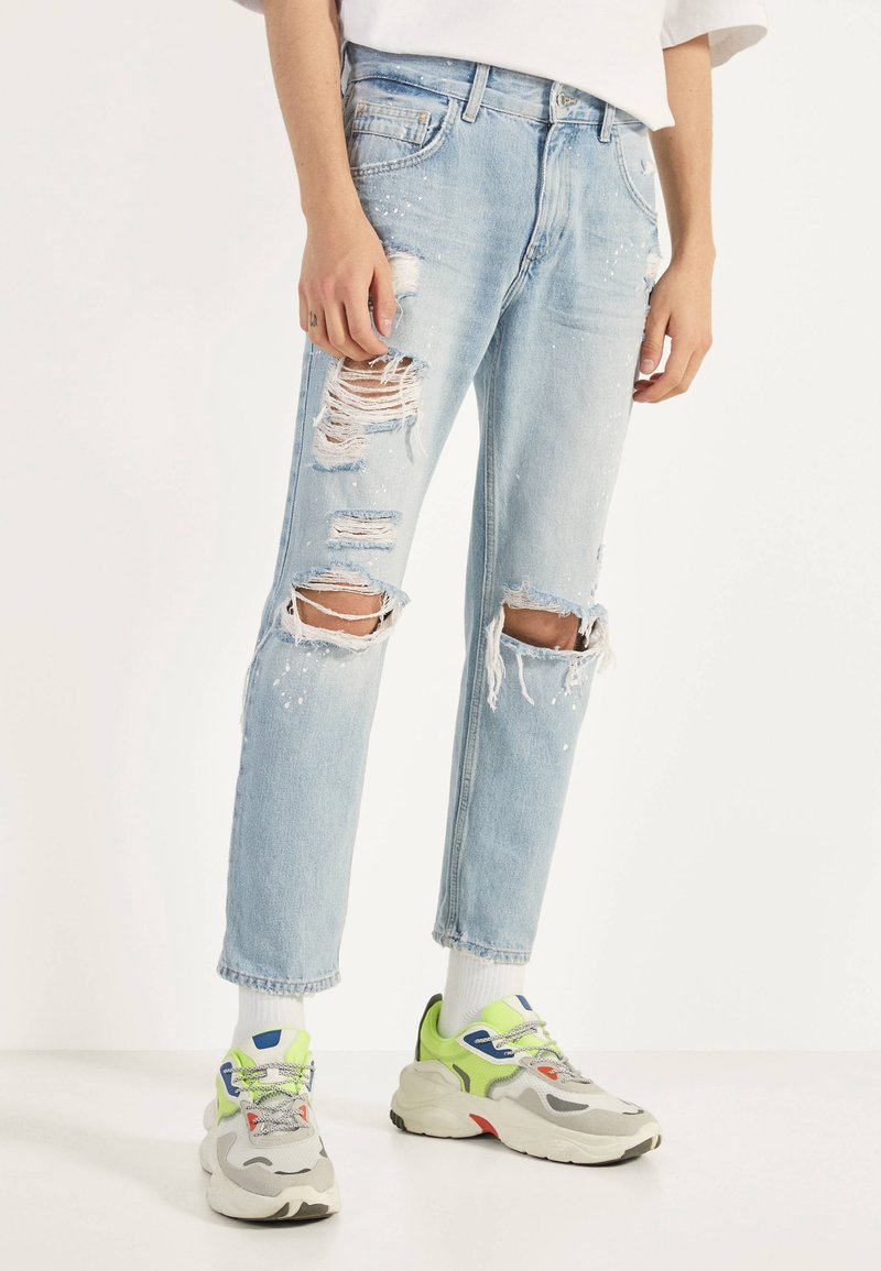 Person som bär ljusblå rivna jeans och flerfärgade kraftiga sneakers med vita strumpor, stående mot en vit bakgrund.