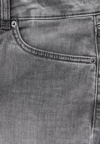 Grauer Denim-Stoff mit Nahaufnahme einer Taschennaht, sichtbaren Nähten und Metallnieten. Glatte Textur mit einem ausgebleichten Erscheinungsbild.