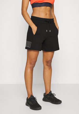 Frau trägt schwarze Puma-Shorts mit Kordelzug, orangefarbenes Croptop und schwarze Sportschuhe, steht mit den Händen in den Taschen.