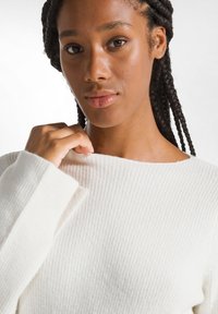 Pulover alb dintr-un tricot cu nervuri, cu decolteu larg și mâneci lungi, evidențiind un material texturat. Mâna modelului trage ușor de decolteu.
