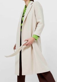 Manteau en laine couleur crème avec une coupe droite, des manches longues et une ceinture nouée. Il possède un col à revers et une finition douce et texturée.