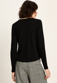 Eine Frau mit kurzen, lockigen Haaren, die ein schwarzes Langarmoberteil und graue Karohosen trägt, steht mit dem Rücken zu einem schlichten Hintergrund.