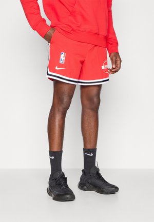 Homme portant un short de basketball rouge NBA Chicago Bulls, des chaussettes noires Nike et des chaussures de sport noires, debout devant un fond blanc uni.