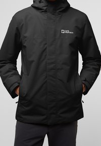 Chaqueta impermeable negra de Jack Wolfskin con capucha, cierre frontal cubierto por una tapeta de tormenta y manos en los bolsillos laterales.