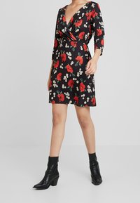 Robe noire portefeuille avec imprimé de coquelicots rouges et de fleurs blanches, décolleté en V, manches trois-quarts, accompagnée de bottines noires.