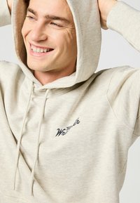 Felpa beige realizzata in tessuto morbido, con tasca a marsupio frontale, cappuccio con coulisse e logo marchiato in nero sul petto.