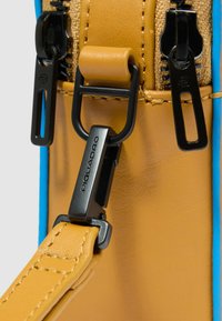 Cinturino in pelle lavorata color cuoio collegato a una fibbia metallica dorata con un dettaglio nero, affiancato da una sezione in tessuto blu e dettagli con cerniera.