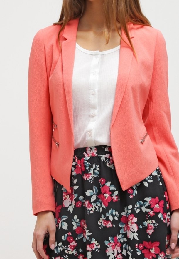 Blazer corail au design structuré, avec des éléments métalliques argentés, porté sur une chemise blanche texturée. Associé à une jupe noire à motif floral.