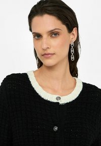 Schwarze Strickjacke mit einem weißen Rippkragen, ausgestattet mit einem dekorativen Knopf und einer strukturierten Oberflächenbeschaffenheit. Das Modell trägt silberne Kettenohrringe.