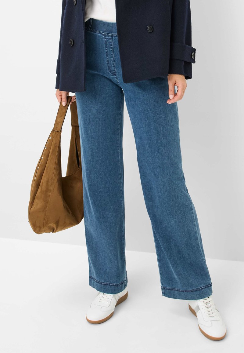 Weite Jeans aus Denim in mittlerem Blauton mit einer subtilen, ausgeblichenen Textur. Kombiniert mit weißen Sneakers und einer braunen Wildleder-Tasche.