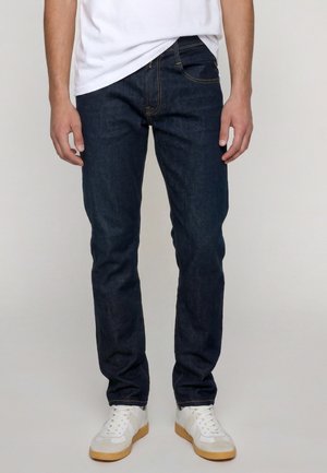 Person iført mørkeblå jeans, hvide sneakers med gummisåler og en hvid t-shirt, der står mod en ensfarvet baggrund.