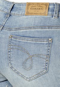Poche arrière d'un jean en denim bleu clair avec des coutures décoratives et un patch de marque en cuir marron clair portant l'inscription "Koroshi Denim Collection Est. 1998."