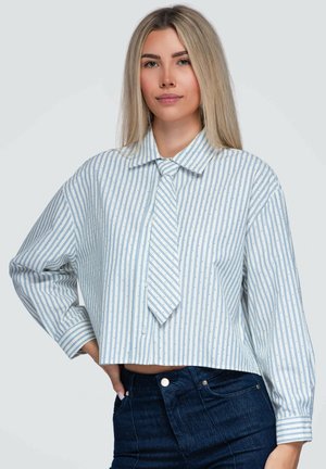 Blondine vrouw die een losse, korte blauw-wit gestreepte blouse draagt met bijpassende stropdas en donkerblauwe spijkerbroek, staande met één hand op de heup.