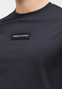 Κοντινό πλάνο μαύρου T-shirt Armani Exchange με ορθογώνιο ταμπελάκι λογότυπου στο στήθος που δείχνει το όνομα της μάρκας με λευκά γράμματα.
