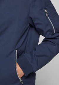 Marinblå jackärm med ribbad mudd, ficka med dragkedja på armen, och hand delvis inne i sidfickan med dragkedja.