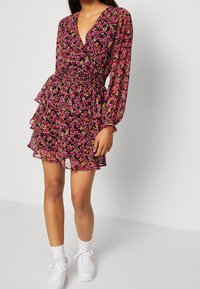 Blommig klänning i mörk tyg med rosa och gula blommor. Har en djup V-ringning, puffärmar och lager av volanger vid kjolen.