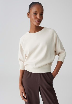Femme à la peau foncée et aux cheveux tirés en arrière, portant un pull ample de couleur crème et un pantalon marron foncé, debout devant un fond gris uni.