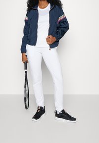 Námořnická větrovka s vícebarevnými pruhy na rukávech, bílý upnutý tričko, bílé tepláky a černé sportovní boty, držící tenisovou raketu.