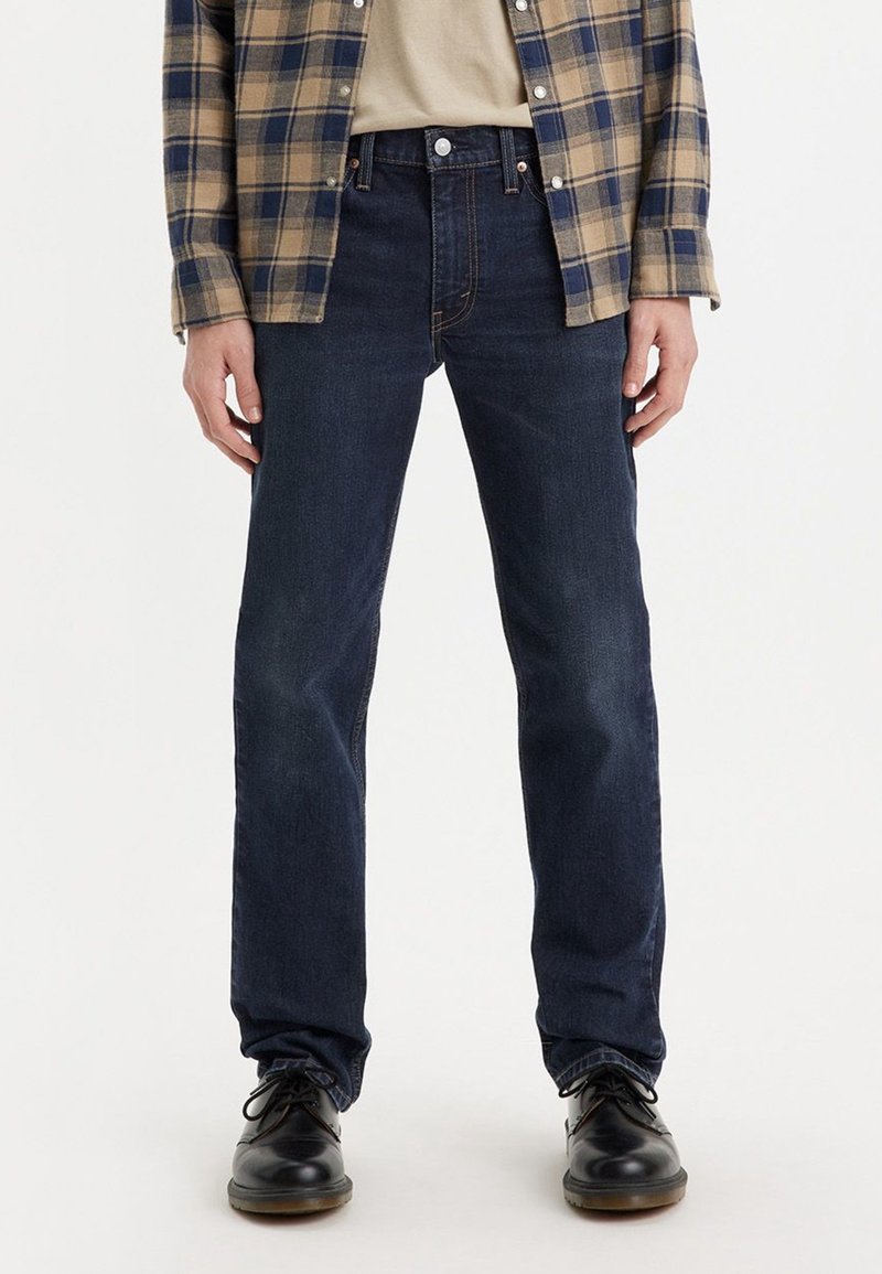 Personne portant un jean droit bleu foncé, des chaussures noires à lacets, une chemise beige et une chemise à carreaux bleu et beige déboutonnée.