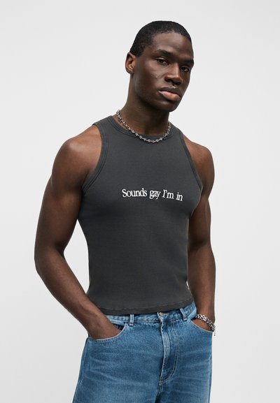 Sivý rebrovaný tielko s bielym textom "Sounds gay I'm in", doplnené modrými džínsami a strieborným retiazkou.