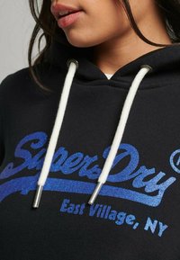 Sudadera negra con un logotipo azul brillante "Superdry" y el texto "East Village, NY", con cordones blancos y puntas metálicas.