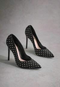 Musta samettiset korkokengät, joissa on terävät varpaat, koristeltuna pienillä valkoisilla niiteillä, pinkki sisävuori ja tyylikkäät stiletto-korkot.