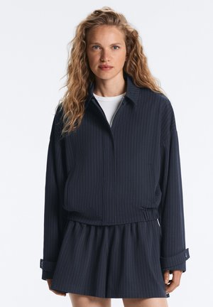 Femme blonde aux cheveux ondulés portant une veste bleu marine foncé à fines rayures et une jupe plissée assortie, debout devant un fond uni.