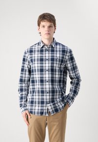 Camisa de cuadros en tonos azul marino y blanco, con botones y mangas largas, que incluye un bolsillo frontal y cuello. Combina con pantalones beige.