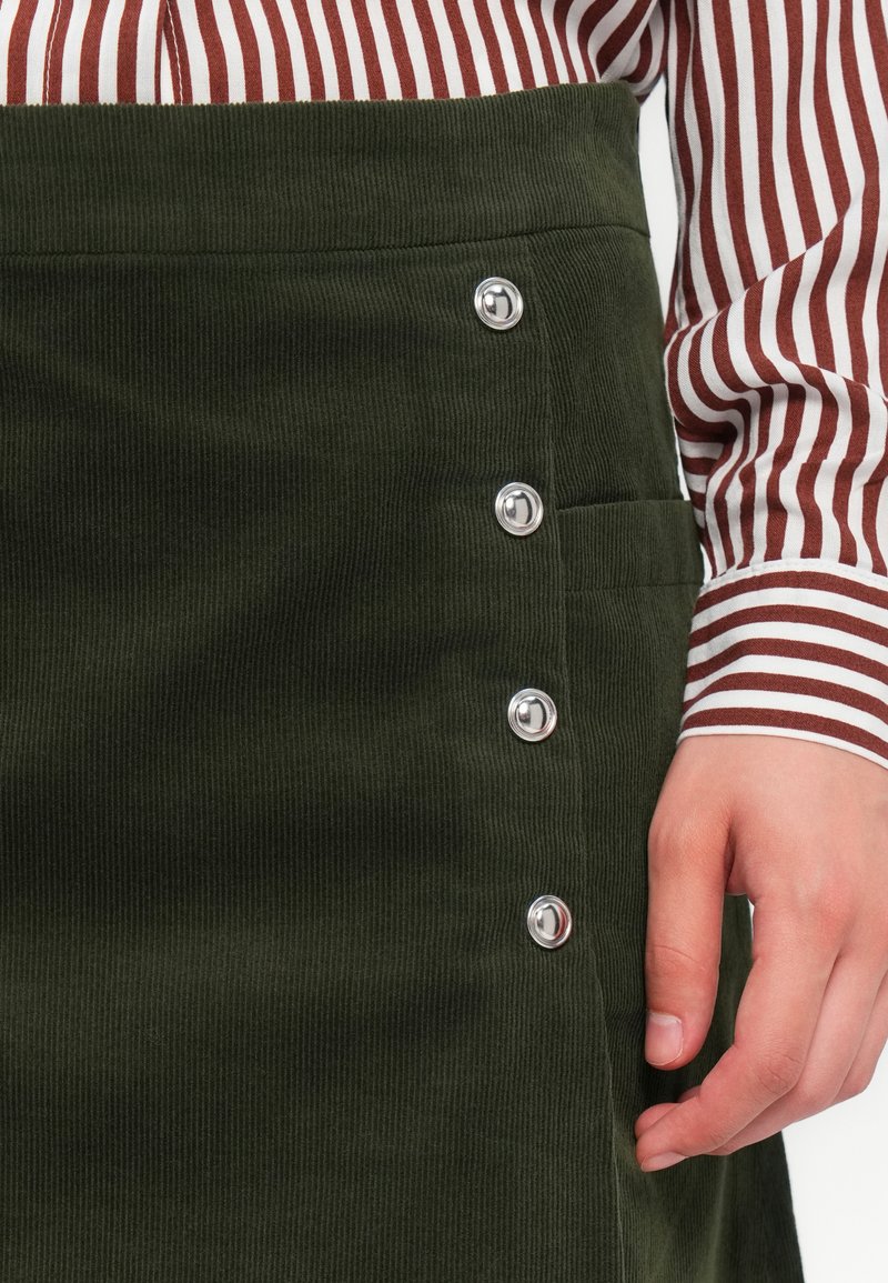 Jupe en velours côtelé vert avec des boutons-pression argentés sur le côté, dotée d'une poche latérale. Tissu rayé d'une chemise visible en arrière-plan.