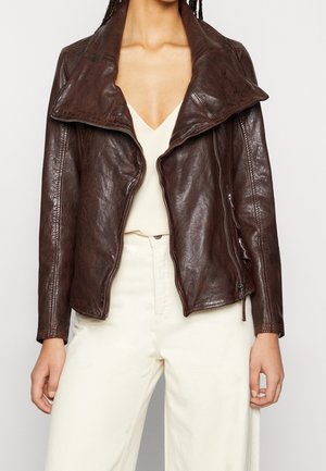 Frau trägt eine offene dunkelbraune Lederjacke über einem beigen V-Ausschnitt-Oberteil und cremefarbenen High-Waist-Hosen, steht vor einem schlichten Hintergrund.