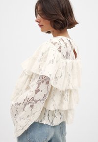 Blouse en dentelle avec des volants superposés, couleur blanc cassé, motif floral, détail du dos ouvert, portée avec un jean bleu clair.