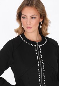 Fekete cipzáras dzseki gyöngy díszítéssel az elején és a nyakkivágásnál, strukturált kialakítással és sima szövet textúrával.