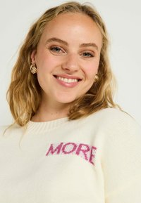 Pull crème avec un col côtelé et "MORE" imprimé en rose, doté d'une texture douce et d'une coupe décontractée. Des boucles d'oreilles dorées ajoutent une touche de finesse.