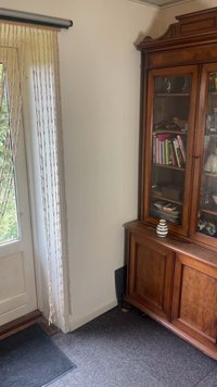 Holzschrank mit Glastüren, der Bücher und Geschirr zeigt, gestreifter Krug darauf, in der Nähe einer Tür mit Perlenvorhang und grauem Fußmatte.