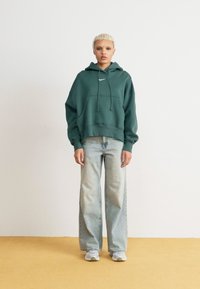 Πράσινη βαμβακερή hoodie με καγκουρό τσέπη, συνδυασμένη με ανοιχτού μπλε φαρδιά τζιν και λευκά αθλητικά παπούτσια. Απλός σχεδιασμός, άνετη εφαρμογή.