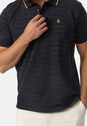 Mand iført mørkegrå poloshirt med orange kant på kraven og ærmerne, beige bukser, justerer kraven med den ene hånd.