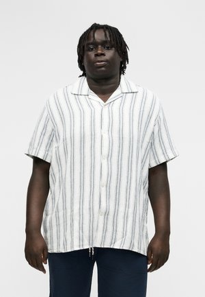 Jaunas vyras dėvi baltą trumpomis rankovėmis marškinius su vertikaliomis juodomis juostelėmis ir tamsias kelnes, stovintis priešais paprastą foną.