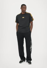 Svart kortermet t-skjorte med gule mønstrede detaljer på ermene og en liten logo. Kombinert med svarte joggebukser med hvit tekst.