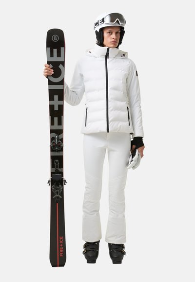 Biała kurtka puchowa z zamykanymi kieszeniami, białe spodnie narciarskie, czarne gogle narciarskie i czarne buty trzymające narty oznaczone napisem "FIRE + ICE."