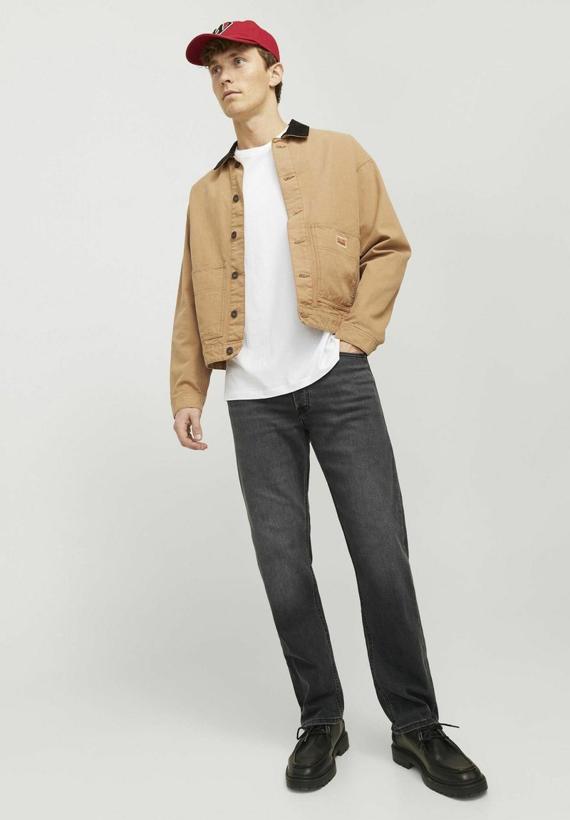 Beige kurz geschnittene Jacke mit schwarzen Kragenakzenten, weißem T-Shirt, dunklen Jeans und schwarzen Schuhen. Das Model posiert vor einem hellgrauen Hintergrund.