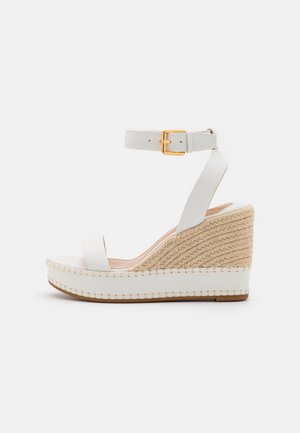Sandale compensée blanche avec talon en jute tressé, large bride avant, bride à la cheville et boucle dorée sur fond blanc.