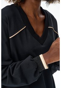 Kobieta ubrana w czarny top z dekoltem w serek, z beżową lamówką na ramionach oraz czarne mankiety z beżowo-białymi paskami, zgięte ramię blisko klatki piersiowej.